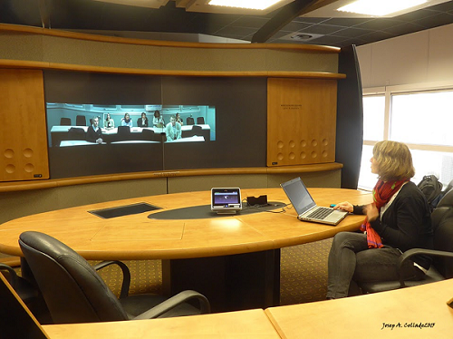 Mesa redonda con una persona viendo una gran pantalla en la que se puede ver una clase con 7 personas