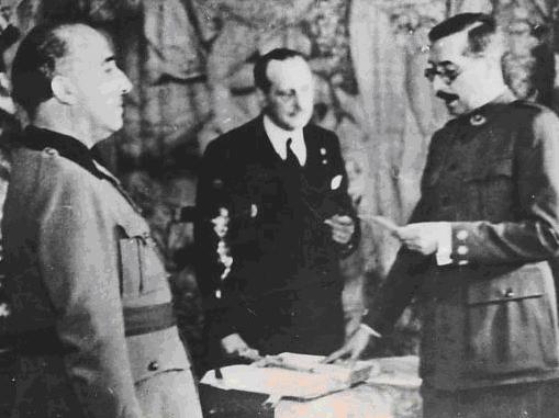 Un militar lee un papel frente a Franco y junto a otra persona en traje civil